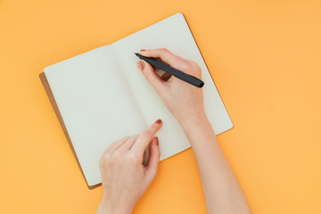 Closeup photo of a woman's hand with a pen, writes in a clean notebook on an orange background, a view from above. Copyspace