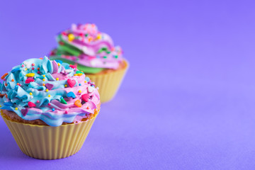 Closeup two colorful cupcakes decorated with sprinkles in shape of hearts on purple background with copy space.