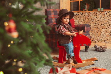 small boy with dark hair, wearing checked red shirt and suede brown vest, hat, is sitting on the...