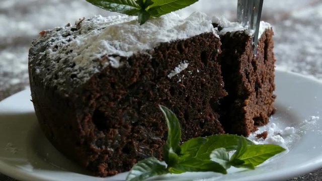 Homemade baked chocolate brownie cake muffled with powdered sugar on a white plate decorated with mint leaves. Fork breaks off piece of brownie pie from the plate