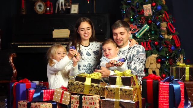 Happy parents shoot festive toy petards with little children in cozy domestic New Year atmosphere under Christmas tree