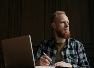 Bearded hipster guy with serious face using laptop computer, writing notes, working project, looking at window and thinking. University student learning language at library, very busy. Brainstorming