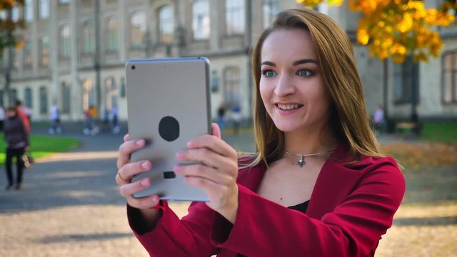 Young attractive female, student having a video call on her tablet, waving with a hand, and showing off her hew hairstyle, outside the university