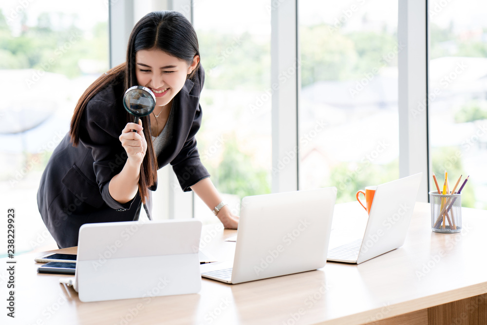 Wall mural asian girl wearing black suit hold magnifying glass looking at the laptop. search in online shopping