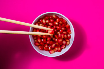 pomegranate seeds with chopsticks in a white bowl on magenta background