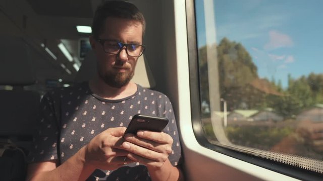 Adult bearded man is typing on touch screen of smartphone, riding in wagon of train in summer. He is turning head and viewing into window