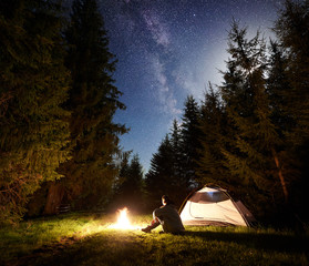 Camping site at night. Tourist tent on forest clearing and male hiker sitting in front of burning campfire under night blue starry sky with Milky way on pine trees background. Outdoor activity concept