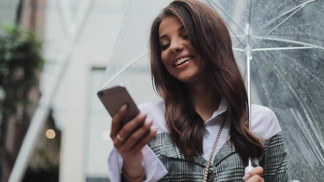 Beautiful young business woman using smartphone on the street in rainy weather, smiling, holding umbrella, communication concept