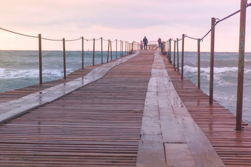 Fototapeta na wymiar Family are walking at the sea pier in storm. Waves splashes through the wooden pier in the sea. Beautiful seascape in sunset.