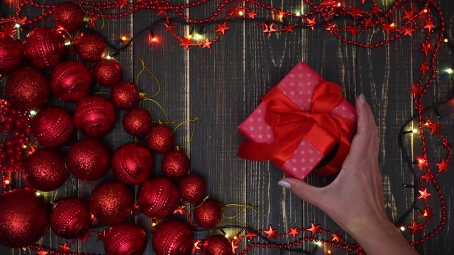 Woman puts small red gift box on brown wooden table. Holiday Christmas background made with many red balls, shine sparkling garland of led lights and wooden painted planks. Top view flat lay 4k video.
