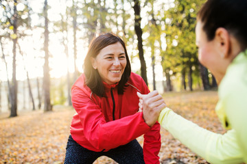 Two female runners stretching outdoors in forest in autumn nature, shaking hands.