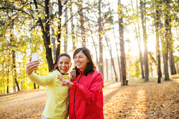 Two female runners with smartphone taking selfie outdoors in forest in autumn nature.