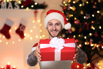 Fototapeta na wymiar Young man in santa hat with gift box near christmas tree at home