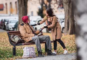 Young woman giving food to homeless beggar man sitting on a bench in city.