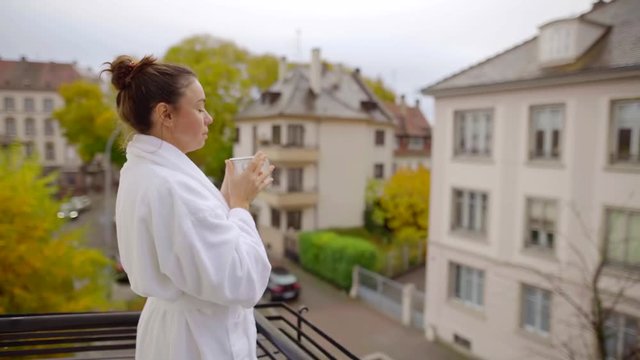 Woman standing on balcony in white robe on balcony with a cup of coffee.