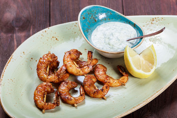 Roasted shrimps on skewers with sauce and lemon on plate on wooden table. Delicious dish of seafood