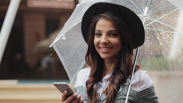Beautiful young business woman using smartphone on the street in rainy weather, smiling, holding umbrella. She looking into the camera. Communication concept