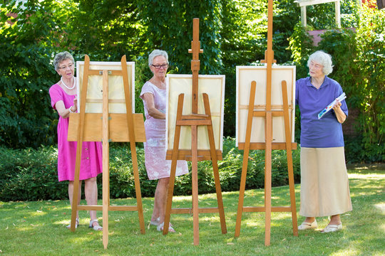 Three Senior Women Painting On Canvas In Garden.