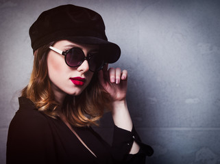 Style redhead girl in black hat and clothes in sunglasses on grey background