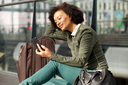 African American Woman Sitting At Station With Suitcase And Mobile Phone