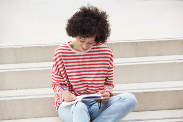 woman with curly hair sitting on steps and writing in diary book