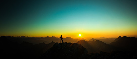 Man reaching summit enjoying freedom and looking towards mountains sunrise.