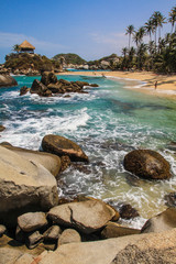 Hochkant Ansicht einer Hütte am Strand mit Palmen in der Karibik mit türkisfarbenem Wasser, Sandstrand und Felsen im Nationalpark Tayrona bei Barranquilla, Kolumbien