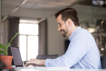 Smiling man using laptop, chatting in social network, typing on keyboard, looking at screen, businessman writing business email, economic report, browsing apps, doing online job, project, development
