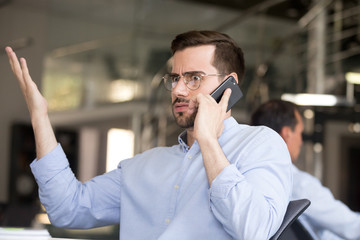 Puzzled businessman with wide eyes and raised hand talking on phone with partner in office at...