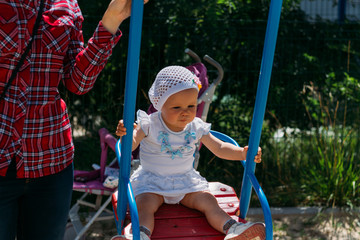 a little girl sits in the arms of a girl, she is hugged behind her back and holding hands.little girl in a white dress and hat riding on a swing, summer sun and heat. playground. childhood serenity