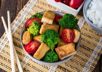 Grilled tofu with broccoli and tomatoes in white bowl on bamboo pad with chopsticks on the left side.