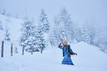 Attractive fashionable slim blond girl in long blue dress, embroidered warm sleeveless fur coat outdoor running in deep clean snow on bright sunny winter day on blurred fir-trees background.
