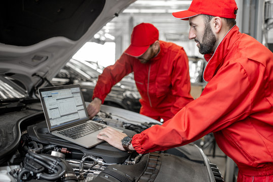 Two auto mechanics in red uniform doing engine diagnostics with computer in the car service