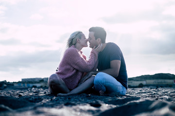 Romantic kiss and love with couple in winter enjoying sit down at the beach in nature outdoor scenic place - millennial people lifestyle concept - defocused ground and attractive young