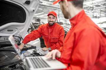 Fototapeta premium Two auto mechanics in red uniform doing engine diagnostics with computer in the car service