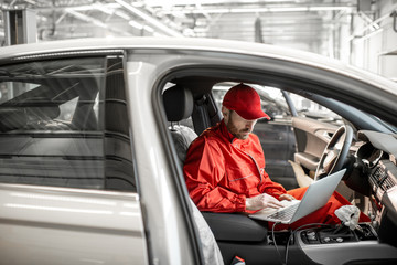Auto mechanic in red uniform diagnosing car with computer sitting on the driver seat at the car...
