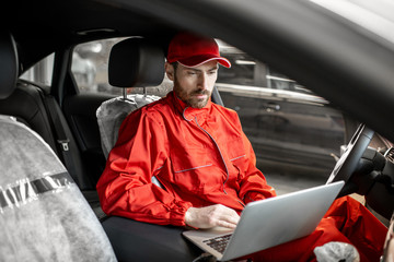Auto mechanic in red uniform diagnosing car with computer sitting on the driver seat at the car...