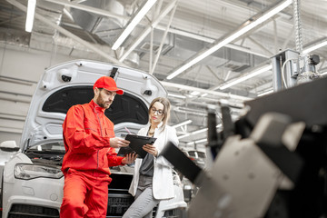 Young woman client with auto mechanic in red uniform standing with some documents at the car...