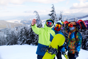 Three snowboarders taking selfie with smartphone camera at ski resort. Friends photographing for social network sharing with snowboards near forest wearing reflective goggles, colorful fashion clothes