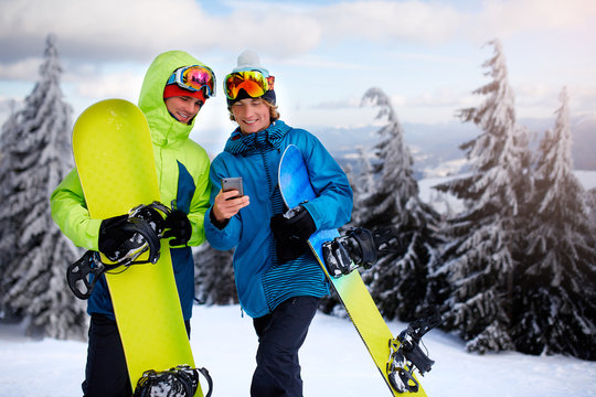 Two snowboarders talking and holding smartphones. Friends looking at cellphone and communicating in social network sharing their photos in app with internet connection wearing colorful fashion outfit.