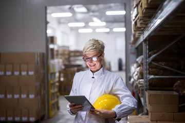 Close up of female factory worker using tablet and holding helmet while standing in warehouse.