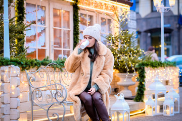 A brunette girl wearing a winter hat and fur coat sitting on a bench and drinks coffee in evening street decorated with beautiful lights at Christmas time