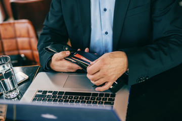 Close up of senior businessman taking credit cart out of wallet while sitting in cafeteria. On the desk laptop and glass of water.
