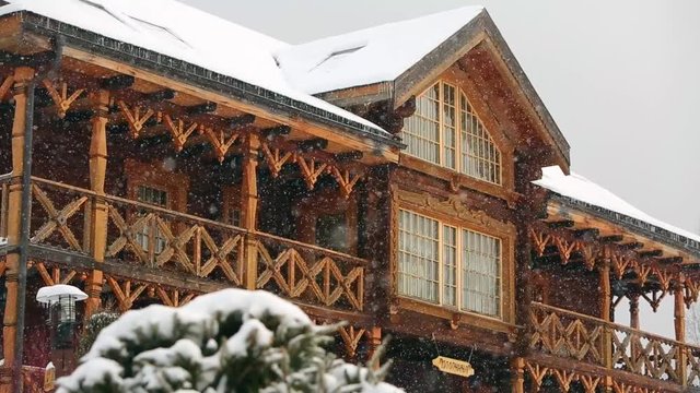 Close view of cottage with mezzanine in mountain village during heavy snowfall with russian style carved wooden facade. Snow falling on log chalets at ski resort. Cold frosty winter day.