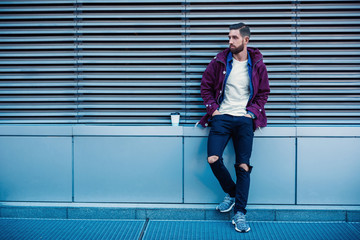 Outdoor fall or winter portrait of handsome hipster man with beard, white t-shirt, blue shirt and maroon jacket holding cup of hot coffee.