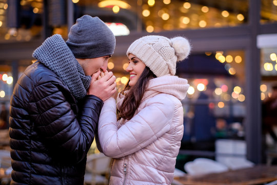 Happy romantic couple wearing warm clothes hold hands and warming while standing in evening street near a cafe outside at Christmas time