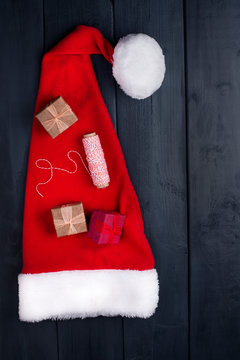 Red Cap with Pompon Santa Claus. On a black wooden background, gifts and Christmas decor. Free space for text. Top view.