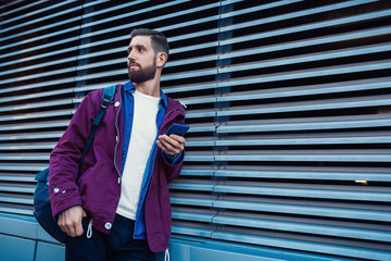 Confident bearded man holding in and mobile phone while standing near street wall in sunny summer...