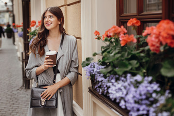 Stylish hipster girl with beautiful hair holding coffee cup and smiling in city street . Gorgeous young woman with paper cup of coffee to-go enjoying time and relaxing in morning