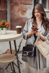 Stylish hipster girl with beautiful hair and smile browsing phone, with coffee cup and bag on wooden table on terrace in city street. Gorgeous happy young woman enjoying time and smiling